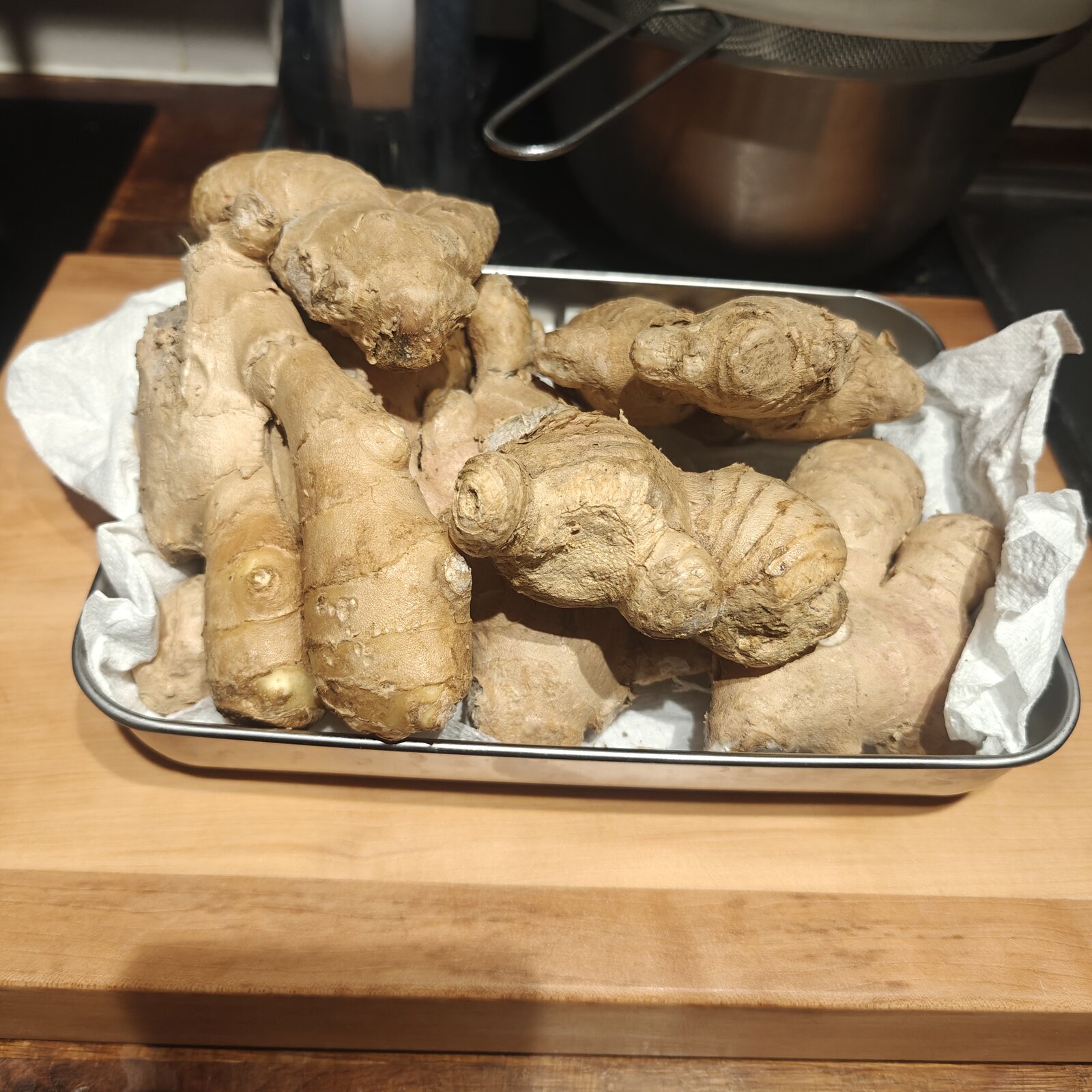 Tray of fresh ginger root knobs on kitchen paper, on a wooden chopping block, with a saucepan and strainer in the background. The 1kg I’m still working through.